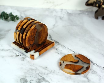 Puede incluir: Un juego de posavasos circulares hechos de rodajas de madera natural y resina blanca translúcida, apilados verticalmente en un soporte cuadrado a juego de madera y resina, con un posavasos circular adicional colocado plano sobre la superficie.