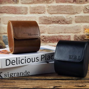May include: Two cylindrical watch cases, one brown and one black, featuring a flap closure. The brown case is embossed with the name Robinson, while the black case is embossed with the initials W.B.L. Both cases have a smooth, structured finish.