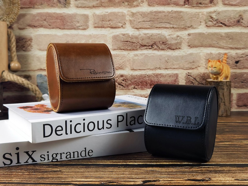 May include: Two cylindrical watch cases, one brown and one black, featuring a flap closure. The brown case is embossed with the name Robinson, while the black case is embossed with the initials W.B.L. Both cases have a smooth, structured finish.