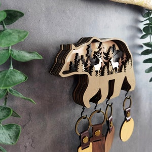 May include: Wooden bear key holder with a layered design featuring a forest scene with deer inside. The bear has five hooks for hanging keys.