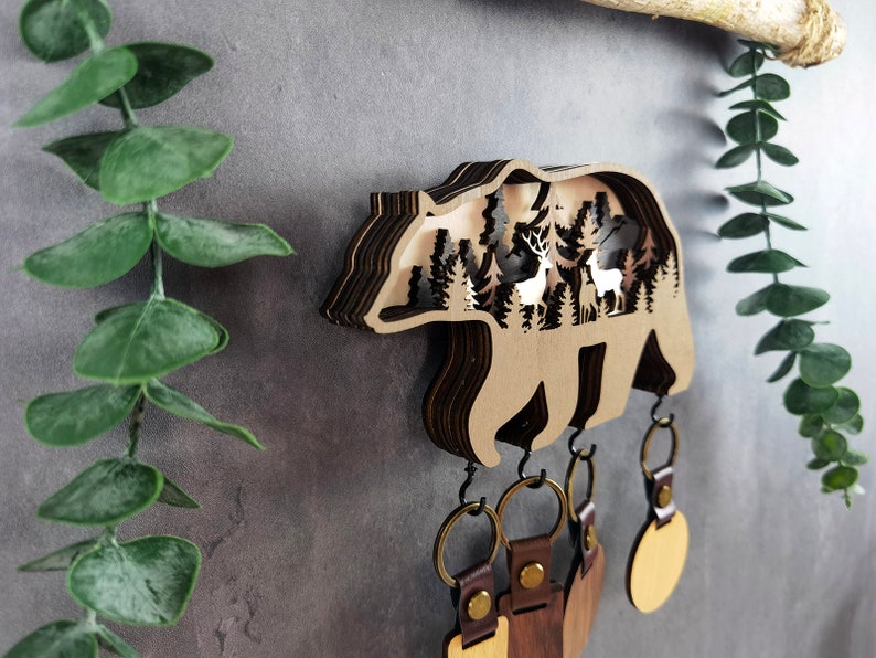 May include: Wooden bear key holder with a layered design featuring a forest scene with deer inside. The bear has five hooks for hanging keys.