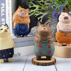 Puede incluir: Cuatro figuras de gatos de madera talladas a mano. Cada gato está pintado con diferentes colores y lleva monos o pantalones cortos. Los gatos se exhiben sobre una superficie blanca con libros y vegetación en el fondo.