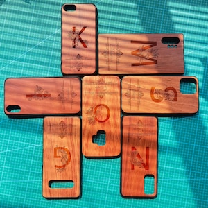 Puede incluir: Nueve fundas de teléfono de madera con nombres e iniciales personalizados. Cada funda presenta un diseño floral único y una letra diferente. Las fundas son todas en tonos marrones y tienen un acabado de madera natural.