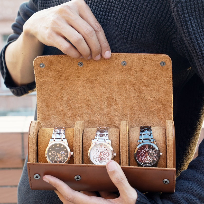 May include: A tan leather watch roll case with a suede interior featuring three individual compartments, each holding a wristwatch. The case has a fold-over flap secured by four metal snap buttons, displaying one black, one white, and one dark metal watch.