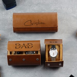 May include: Three brown cylindrical watch cases with snap closures. One closed case is engraved with the name Christian. Another open case displays the word DAD with names, and the third open case shows K B and a silver wristwatch.