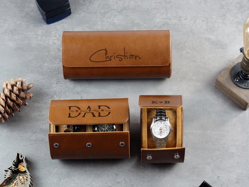 May include: Three brown cylindrical watch cases with snap closures. One closed case is engraved with the name Christian. Another open case displays the word DAD with names, and the third open case shows K B and a silver wristwatch.