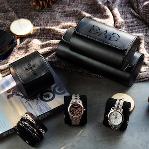 May include: A black leather watch roll with the word DAD embossed on the lid, a smaller black leather watch case with an embossed cross, two wristwatches on black cushions, and a stack of bracelets on a black cushion, all arranged together.