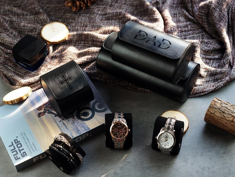 May include: A black leather watch roll with the word DAD embossed on the lid, a smaller black leather watch case with an embossed cross, two wristwatches on black cushions, and a stack of bracelets on a black cushion, all arranged together.