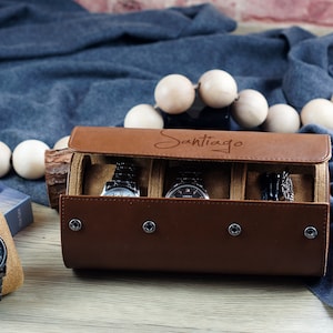May include: A brown leather watch roll case with the name Santiago engraved on the lid, featuring four silver snap buttons and an interior lined with tan fabric. The case holds two wristwatches, while a third black metal watch sits on a tan pillow.