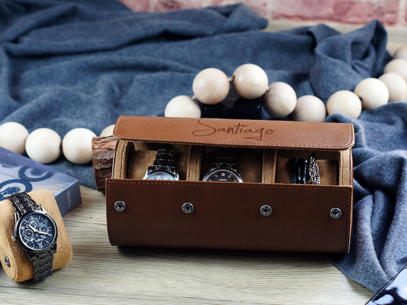 May include: A brown leather watch roll case with the name Santiago engraved on the lid, featuring four silver snap buttons and an interior lined with tan fabric. The case holds two wristwatches, while a third black metal watch sits on a tan pillow.