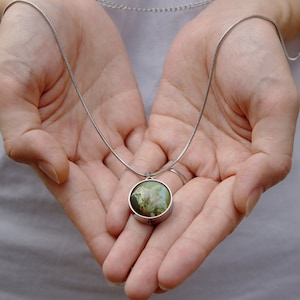 Op de afbeelding: Een zilveren ketting met een ronde hanger met een groen en wit bloemenmotief.