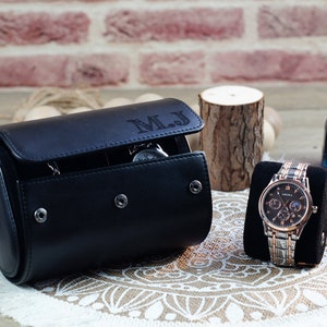 May include: A black cylindrical watch roll case with the initials M.J. engraved on the flap, featuring three silver snap buttons. Beside it, a silver and rose gold wristwatch with a dark face is displayed on a black velvet cushion.