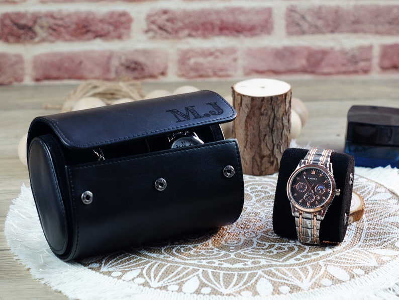 May include: A black cylindrical watch roll case with the initials M.J. engraved on the flap, featuring three silver snap buttons. Beside it, a silver and rose gold wristwatch with a dark face is displayed on a black velvet cushion.