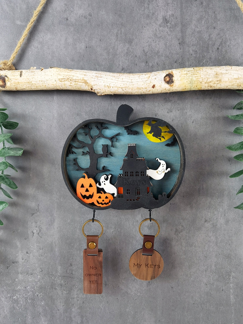 May include: A wooden Halloween-themed key holder shaped like a pumpkin with a haunted house, ghosts, and a witch flying on a broomstick. The key holder has two key rings with tags that say "No Owner Yet" and "My Keys".
