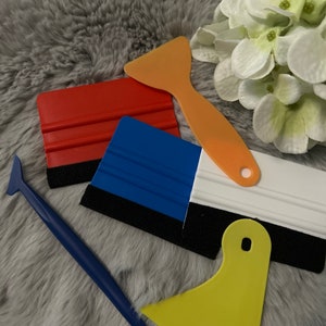 Könnte beinhalten: Eine Sammlung farbiger Rakel in Rot, Blau, Orange, Gelb und Weiß. Die Rakel haben schwarze Filzkanten und sind auf einer grauen, strukturierten Oberfläche angeordnet. Einige haben einen Griff, andere ein flaches Design. Die Werkzeuge dienen zum Aufbringen von Vinyl.