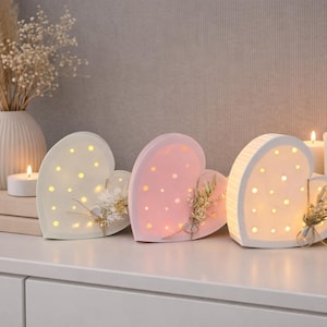 Op de afbeelding: Drie hartvormige decoratieve lichten in crème, roze en wit, elk met kleine, warme lichten. De harten zijn versierd met gedroogde bloemenaccenten. Kaarsen en een vaas met gedroogde bloemen zijn ook aanwezig.