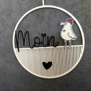 Könnte beinhalten: Ein kreisförmiger Wandbehang aus Holz mit dem Wort Moin in schwarzer Schreibschrift, einer weißen Möwe mit roter Strickmütze und einem kleinen schwarzen Herz. Die untere Hälfte des Kreises hat vertikale Lamellen und ein mittiges Herz.