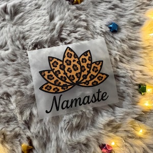 Könnte beinhalten: Ein quadratischer, transparenter Aufkleber mit einem Lotusblumen-Design im Leopardenmuster. Das Wort "Namaste" ist in einer kursiven Schrift unter der Blume geschrieben. Der Aufkleber befindet sich auf einem grauen, flauschigen Hintergrund.