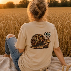 Könnte beinhalten: Ein beiges kurzärmeliges T-Shirt mit einem Grafikdruck auf der Rückseite, der eine Cartoon-Schnecke mit einem braun-schwarzen Leopardenmuster-Gehäuse, großen grünen Augen und einem lächelnden Gesicht zeigt, kombiniert mit einer blauen Jeans mit Riss.