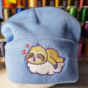 May include: Light blue knit beanie featuring an embroidered sleeping sloth on a cloud. The sloth is brown and yellow with purple accents. The beanie is made of a soft, ribbed knit material. A fun and cozy accessory.