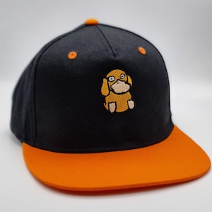 May include: Black and orange baseball cap with an embroidered cartoon dog on the front. The cap has a flat brim and a button on top. The dog is light brown with black outlines and a white face.