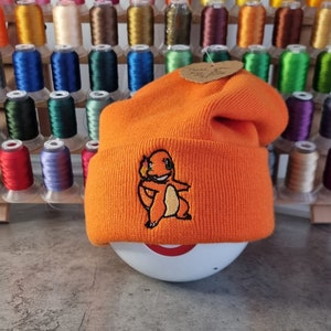 May include: Orange beanie with a small embroidered cartoon character. The beanie is sitting on a white surface.