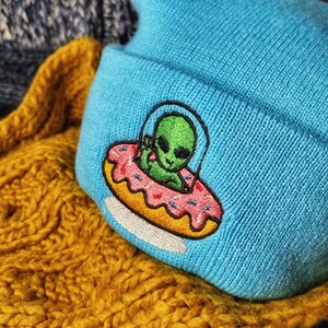 Peut inclure: Un bonnet en tricot bleu avec un extraterrestre brodé chevauchant un vaisseau spatial en forme de beignet. Le bonnet est posé sur un tissu en tricot jaune et bleu.