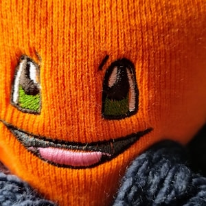 May include: Orange knitted plush toy with a smiling face, green eyes, and a pink tongue sticking out. The toy is wearing a blue knitted hat.