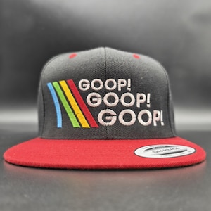May include: A black snapback hat with a red brim. The hat features the embroidered text "GOOP! GOOP! GOOP!" in white, with a colorful rainbow design. The hat is a classic, flat-brim style.