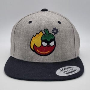 May include: A grey and black snapback cap with an embroidered cartoon chilli pepper with flames. The chilli pepper has a red body, green stem, and angry eyes. The cap has a flat peak and a snap closure.