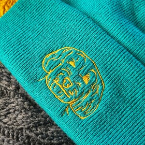 Könnte beinhalten: Eine tealblaue Strickmütze mit einem gelben gestickten Gesichtsmotiv. Die Mütze ist umgeklappt und liegt auf einem grauen Strickstoff.