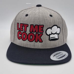 May include: A grey snapback cap with a navy blue peak. The cap displays the text "LET ME COOK" in red block letters and a white chef's hat graphic. It has a flat peak and a snap closure.
