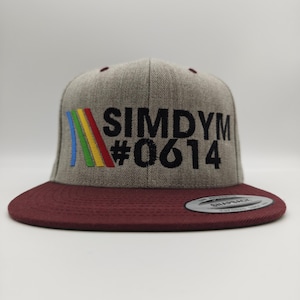 May include: A gray flat-brimmed snapback hat with a maroon brim. The hat features the text "SIMDYM #0614" in black, with a colorful rainbow design. The hat is a stylish accessory.