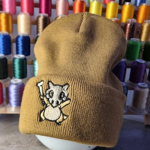 May include: A tan beanie hat with a Cubone embroidered patch. The hat is made of a knitted material and has a folded brim. The background features spools of colourful thread, suggesting a craft or handmade item.
