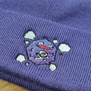 May include: Purple beanie with an embroidered image of a purple and white cartoon character with a skull and crossbones. The character is surrounded by white clouds.