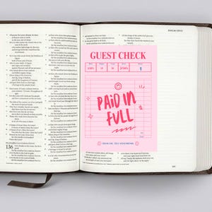 May include: A pink guest check with the words "PAID IN FULL" in a handwritten style. The check is open inside a Bible. The check also has the words "GUEST CHECK" at the top.