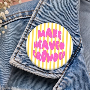 May include: A circular pinback button featuring a white and yellow vertical striped background with the text "MAKE HEAVEN CROWDED" written in a bold, rounded, bubble-style pink font. The button is attached to the collar of a blue denim jacket.