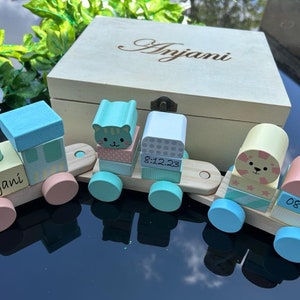 May include: A wooden toy train with four cars. The first car is a light blue and pink engine with the name "Anjani" on it. The second car is light blue and pink with a cat face on it. The third car is light blue and white with the date "8.12.23" on it. The fourth car is light blue and pink with a bear face on it and the time "08:54" on it. The train is in a wooden box with the name "Anjani" engraved on it.