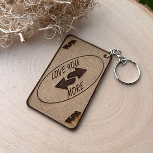 May include: A wooden keychain with the text "LOVE YOU MORE" and two arrows. The keychain has a silver keyring. The background is a light-coloured wooden surface with dried plant material. The keychain is rectangular with rounded corners.