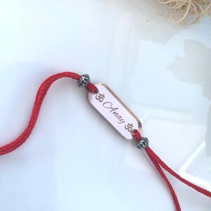 May include: A red cord bracelet with a rectangular, light pink charm. The charm has the name "Anay" and an Om symbol engraved on it. The bracelet also features decorative silver beads.