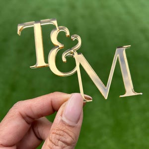May include: A gold-coloured acrylic monogram displaying the letters "T & M". The letters are in a decorative font, with a reflective finish. The monogram is held against a green background, possibly for outdoor use or a natural setting.