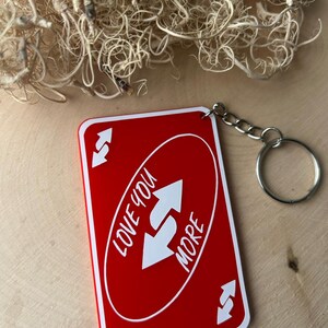 May include: A red keychain with the text "Love You More" in white, featuring a reverse card design. The keychain has a silver chain and ring. The background is a light wood surface.