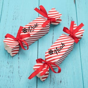 Puede incluir: Dos cajas de cracker de Navidad a rayas rojas y blancas con lazos de cinta roja. Las cajas están decoradas con un copo de nieve y los nombres "Rose" y "Noah".