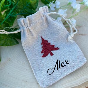 May include: A small, off-white fabric drawstring bag with a red glitter Christmas tree and the name "Alex" written in black cursive.