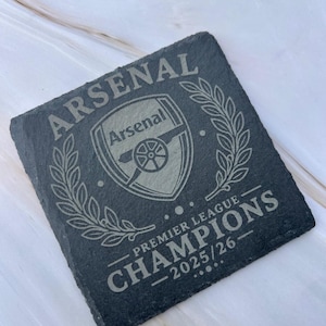 May include: A square dark grey slate coaster featuring an engraved Arsenal Football Club crest at the centre, flanked by laurel wreaths. The text reads ARSENAL at the top and PREMIER LEAGUE CHAMPIONS 2025/26 at the bottom in a bold, serif font.