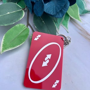 May include: A rectangular red keyring featuring a white oval in the centre with two opposing white arrows inside. The card design includes two additional white arrows in the top left and bottom right corners, attached to a silver metal ring.