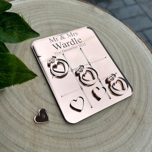 May include: Rose gold mirrored game board with the text "Mr & Mrs Wardle 31st December 2022" and heart-shaped cutouts. The board is on a wooden surface.