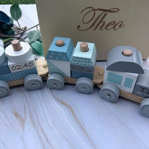 May include: A wooden toy train with three connected carriages in shades of blue, grey, and white. The train features stacking blocks, pegs, and wheels. The text 02:40, 11.09.26, and Theo are printed on the blocks. A wooden box with the name Theo is behind.