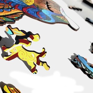 Puede incluir: Piezas de rompecabezas de madera en varias formas y colores. Las piezas presentan tonos amarillos, rojos, azules y marrones. Una pieza tiene la forma de un ciervo corriendo. Las piezas del rompecabezas est&aacute;n sobre un fondo blanco.