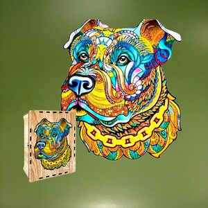 Puede incluir: Un rompecabezas de madera de un perro colorido con una caja a juego. La imagen del perro es una vibrante mezcla de amarillo, azul, naranja y verde. El rompecabezas y la caja est&aacute;n sobre un fondo verde.
